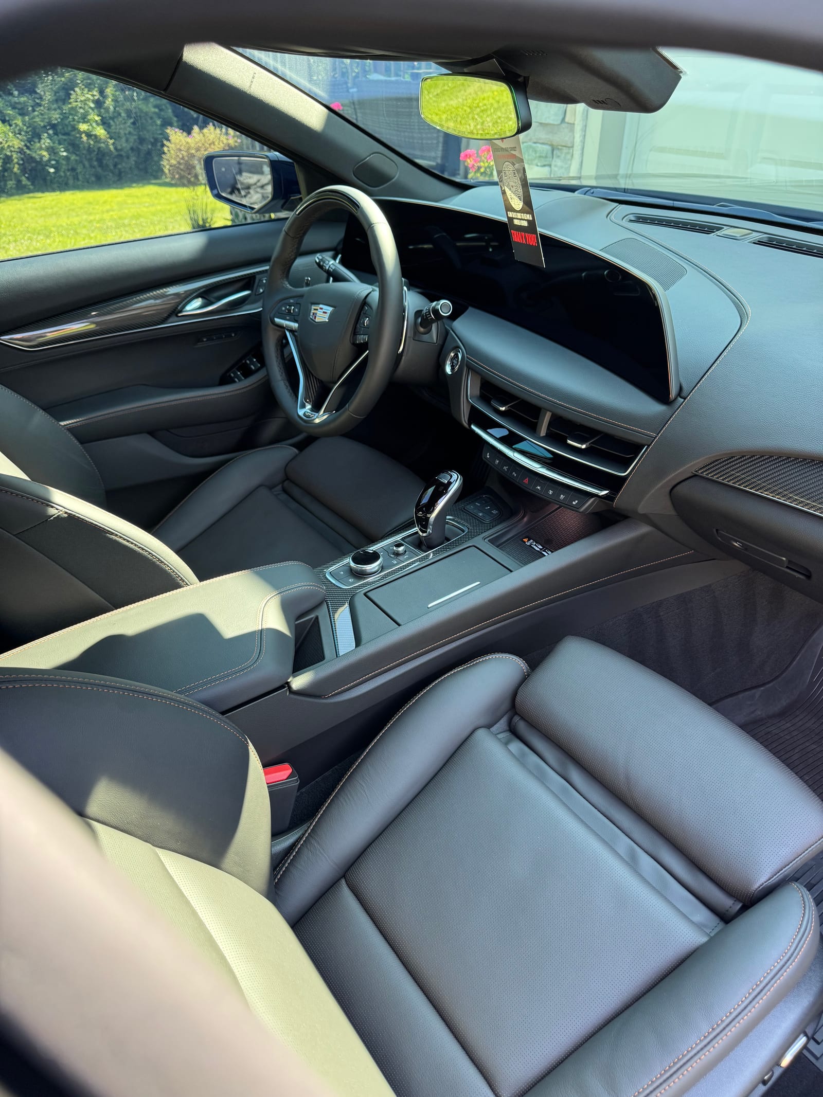 Freshly detailed Cadillac interior — black leather seats with contrast stitching, carbon-fiber trim, clean center console and dashboard — shot from the passenger side in a Mid-Michigan driveway, greenery visible through the windows.