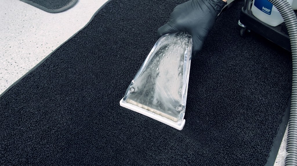Gloved After Hours detailer drawing a hot-water extraction wand across a black car floor mat, water lifting up through the clear wand head — deep extraction that pulls ground-in dirt and salt out of the carpet fibers, not just the surface.