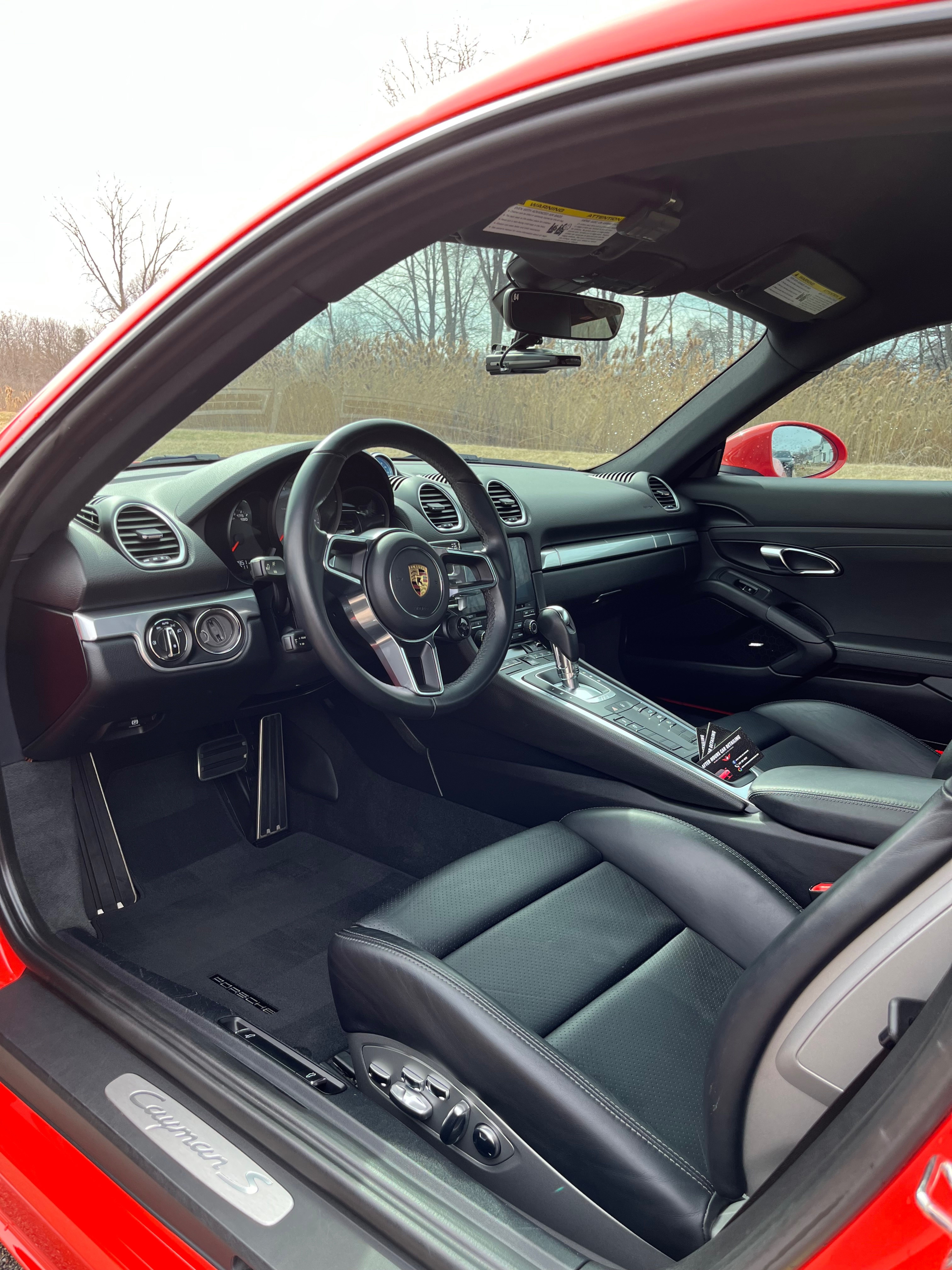 Spotless red Porsche 718 Cayman S interior — black leather seats, detailed dashboard, and an After Hours Car Detailing business card placed on the console. The kind of interior Mid-Michigan customers book online for.